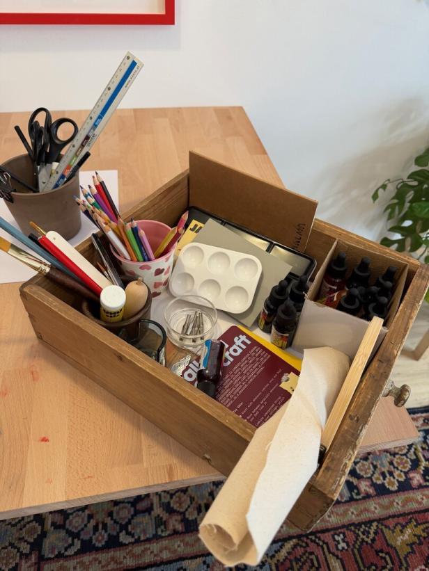 Eine Holzkiste mit Bastelmaterialien wie Stiften, Pinseln, Farben, Schere, Lineal und Papierrolle steht auf einem Tisch.