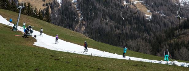 Skifahrer und eine Schneekanone auf einer Skipiste mit wenig Schnee.