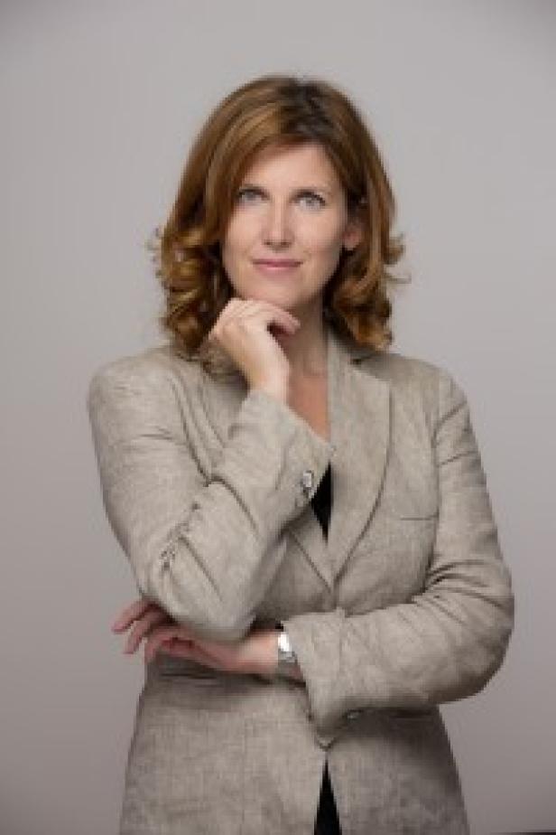 Frau mit schulterlangen, lockigen Haaren trägt einen beigen Blazer und blickt nachdenklich nach vorn.