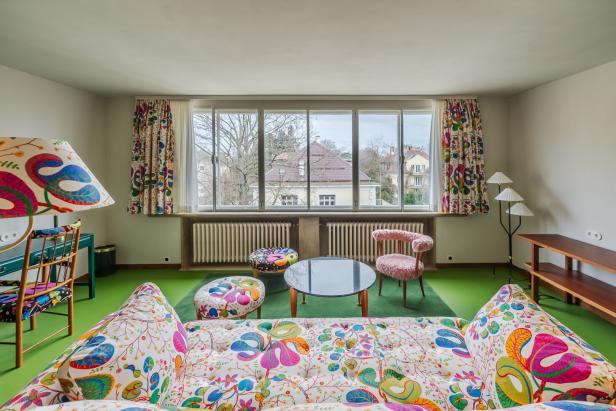 Wohnzimmer mit großen Fenstern, bunten Möbeln und Vorhängen, grünem Boden und Blick auf Häuser und Bäume draußen.