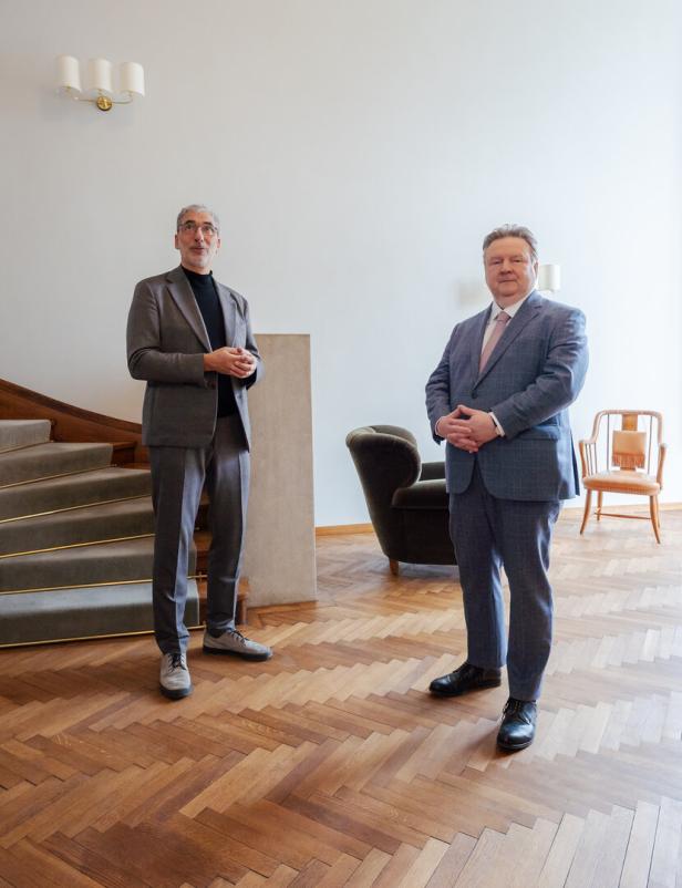 Zwei Männer in Anzügen stehen in einem elegant eingerichteten Raum mit Holzfußboden und Treppe.