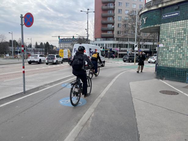 Zwei Radfahrer fahren auf einem markierten Radweg an einer belebten Kreuzung in der Stadt entlang.