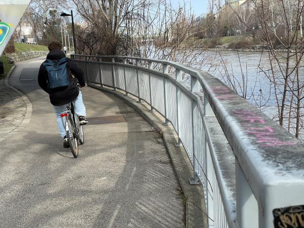 Eine Person mit Rucksack fährt auf einem Fahrradweg am Fluss entlang, umgeben von kahlen Bäumen.