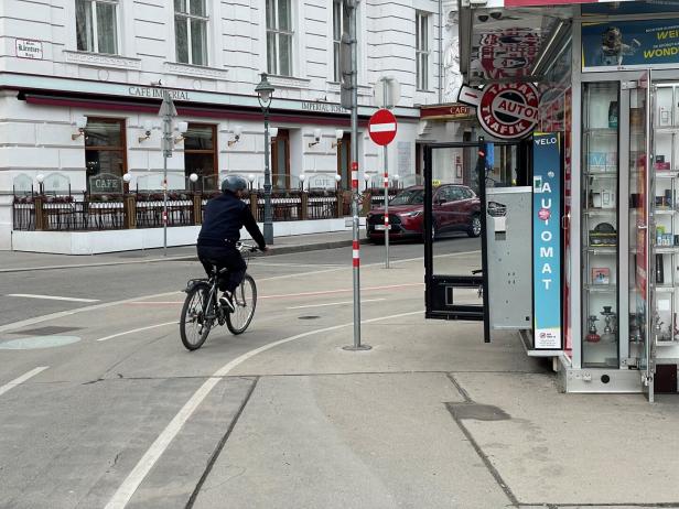 Eine Person fährt mit dem Fahrrad auf einer Straße an einem Tabakautomaten und einem Café vorbei.