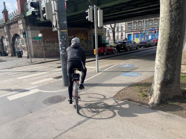 Eine Person mit Helm fährt auf einem Fahrradweg unter einer Brücke, während Autos auf der Straße daneben stehen.