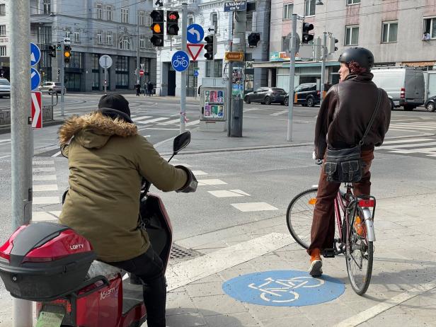 Ein Radfahrer und ein Motorrollerfahrer warten an einer Kreuzung auf grüner Ampel, umgeben von Verkehrszeichen.