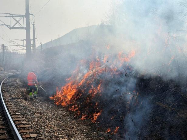 Ein Arbeiter in Schutzkleidung kontrolliert ein Feuer neben Bahngleisen, Rauch steigt auf.