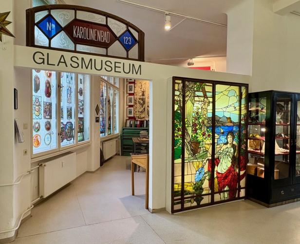 Eingangsbereich eines Glasmuseums mit buntem Jugendstilfenster und Vitrinen mit Ausstellungsstücken.