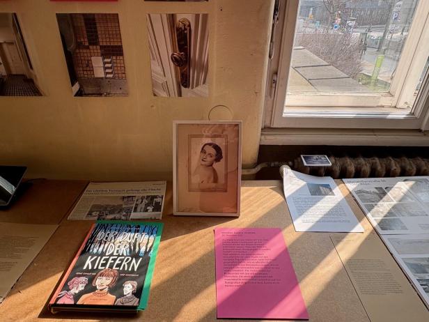 Auf einem Tisch liegen Bücher, Fotos, Zeitungsartikel und Dokumente, im Hintergrund ist ein Fenster mit Blick nach draußen.