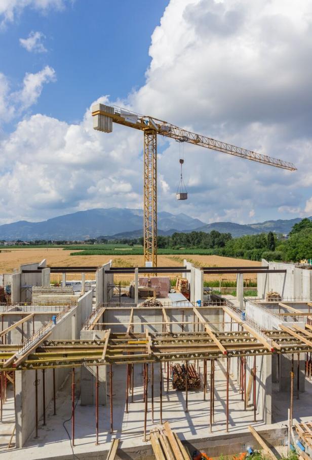 Ein gelber Baukran hebt Baumaterial über eine teilweise errichtete Betonbaustelle auf dem Land.