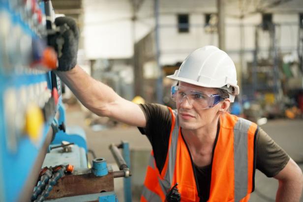 Eine Person mit Schutzhelm und Warnweste bedient konzentriert eine Maschine in einer Werkhalle.