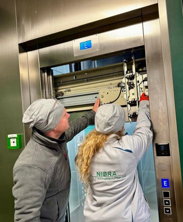Zwei NIBRA-Mitarbeiter reparieren einen Aufzug mit geöffneter Tür und sichtbarer Mechanik.