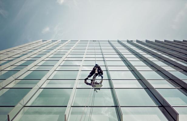 Eine Person reinigt mit Seiltechnik die Glasfassade eines hohen modernen Gebäudes.