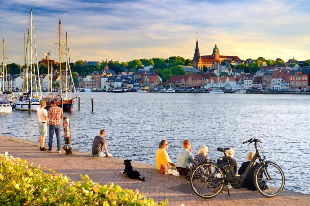 Menschen sitzen am Ufer mit Blick auf einen historischen Stadtkern und Segelboote im Hafen.
