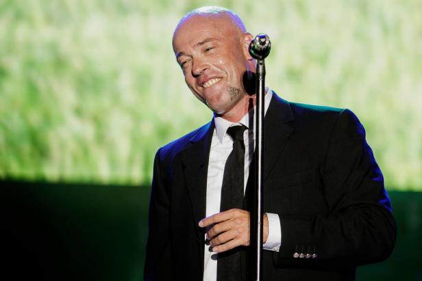 Der Graf von Unheilig steht im schwarzen Anzug und mit Mikrofon auf einer Bühne und lächelt während eines Auftritts.