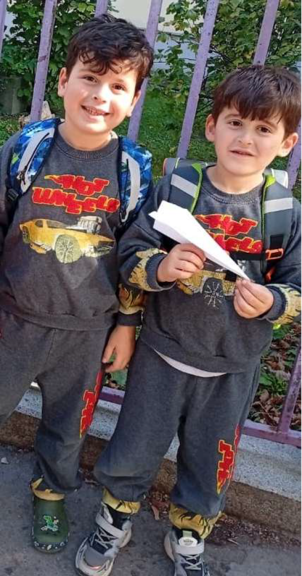 Zwei Burschen im gleichen Outfit stehen vor einem Zaun und lächeln in die Kamera - einer hält einen Papierflieger.