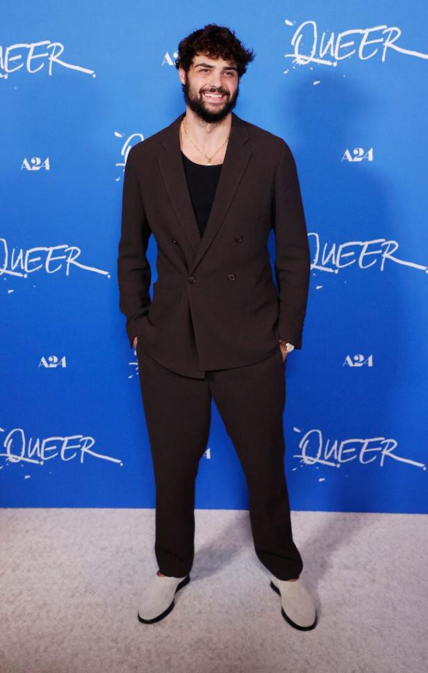 Ein Mann im braunen Anzug steht vor einem blauen Hintergrund mit dem Schriftzug "Queer".