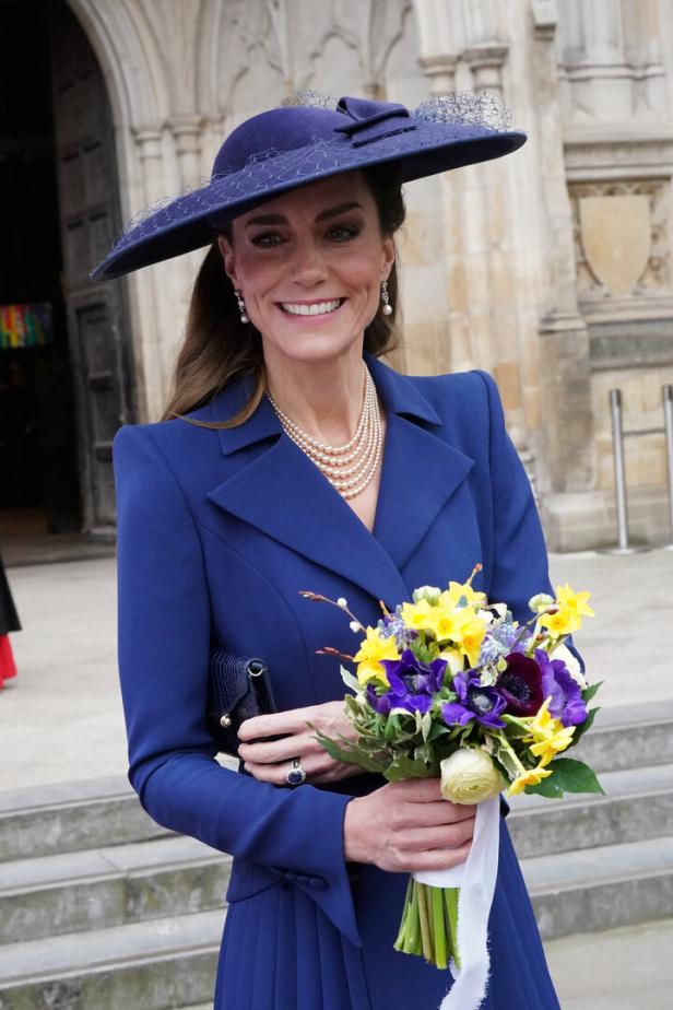Kate trägt einen blauen Hut und Blazer und hält einen Blumenstrauß.