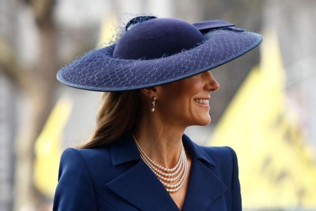 Kate mit elegantem dunkelblauem Hut, Perlenkette und Ohrringen lächelt und trägt ein passendes Jackett.
