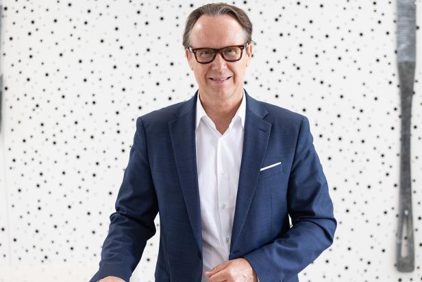 Ein Mann mit Brille und blauem Sakko lächelt vor einem weiß gepunkteten Hintergrund.