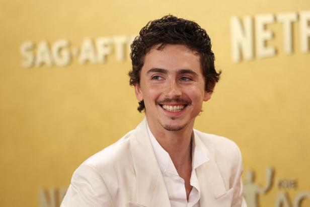 Ein lächelnder Timothée Chalamet mit dunklen Locken und weißem Anzug vor goldenem Hintergrund.