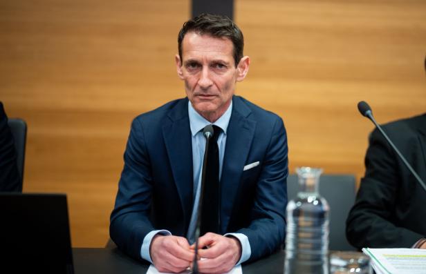 Ein Mann im Anzug sitzt an einem Tisch mit Mikrofon und Wasserflasche vor holzvertäfeltem Hintergrund.