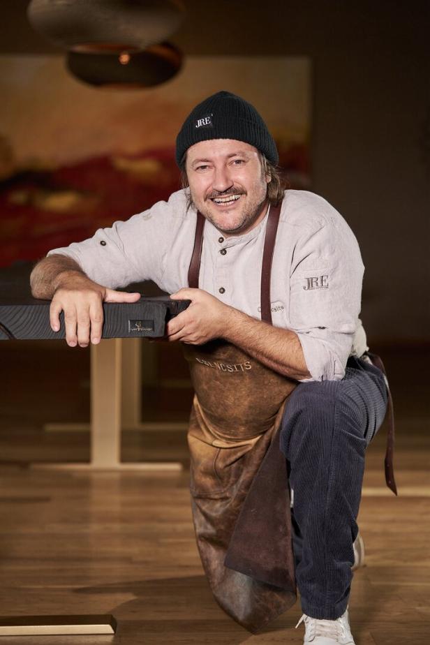 Ein Mann mit Schürze und Mütze kniet lachend an einem Tisch in einem warm beleuchteten Raum.