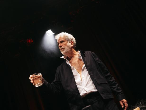 Ein älterer Mann mit grauem Haar und Bart steht im Scheinwerferlicht und hält ein Glas in der Hand.