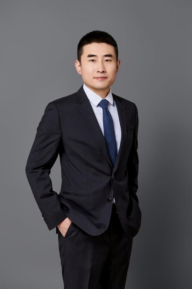 Ein Mann im dunklen Anzug mit weißem Hemd und blauer Krawatte steht vor grauem Hintergrund.