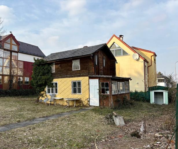 Ein kleines, älteres Haus mit gelber Fassade und Holzaufbau steht auf einem verwilderten Grundstück neben modernen Gebäuden.
