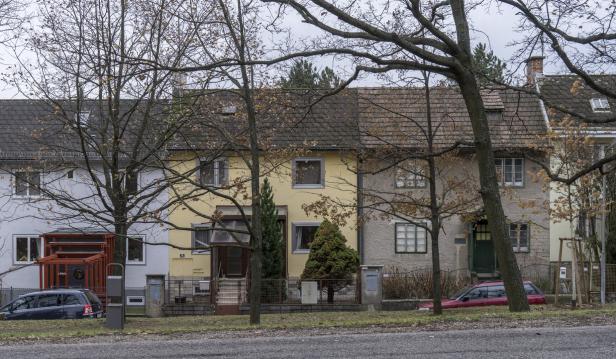 Mehrere Reihenhäuser mit Vorgärten und kahlen Bäumen stehen an einer Straße, davor sind zwei geparkte Autos zu sehen.