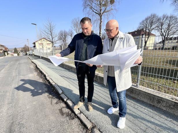 Zwei Männer stehen auf einem Gehweg neben einer Straße und betrachten gemeinsam große Baupläne.