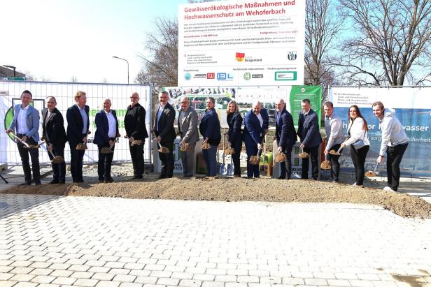 Vierzehn Personen stehen nebeneinander und heben mit Schaufeln Erde auf einer Baustelle aus.