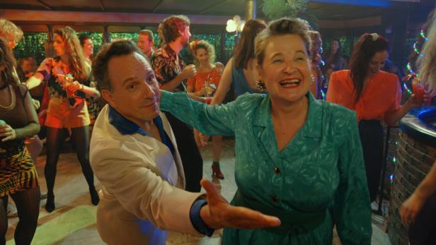 Ein Mann und eine Frau tanzen lachend auf einer bunten Party mit vielen weiteren feiernden Menschen im Hintergrund.