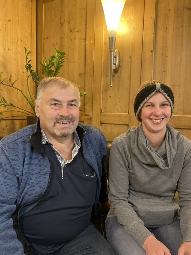 Ein älterer Mann und eine junge Frau sitzen nebeneinander vor einer holzvertäfelten Wand.