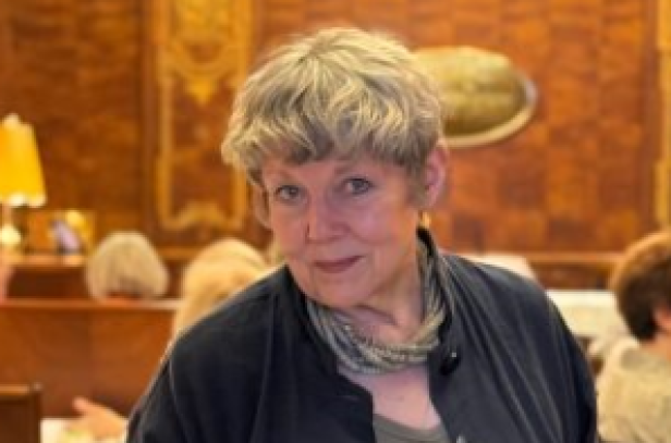 Ältere Frau mit grauen Haaren in einem holzvertäfelten Raum mit mehreren Personen im Hintergrund.