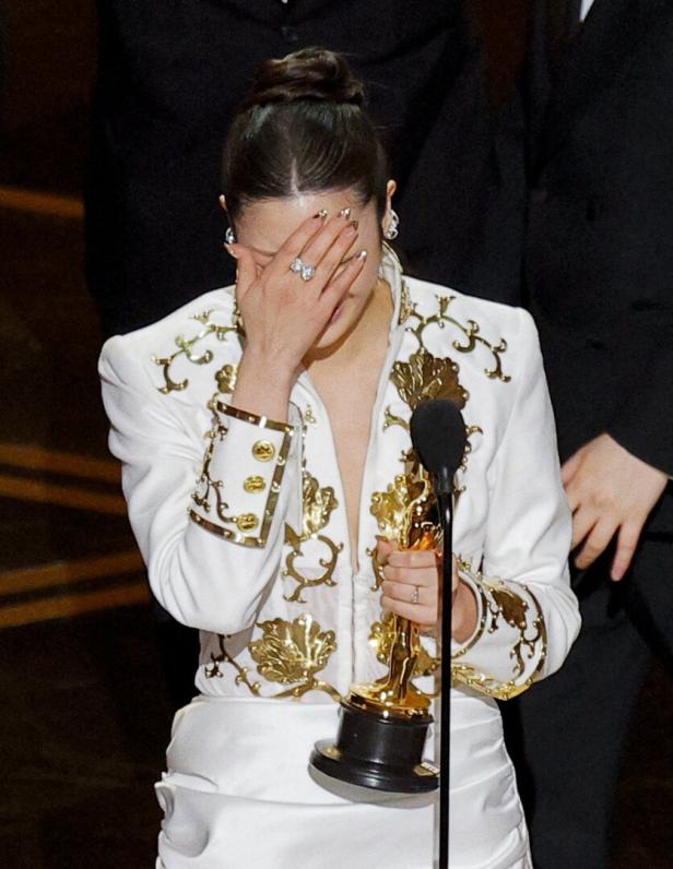 Sängerin Ejae in weiß-goldenem Anzug hält eine Trophäe und bedeckt ihr Gesicht mit der Hand vor einem Mikrofon.