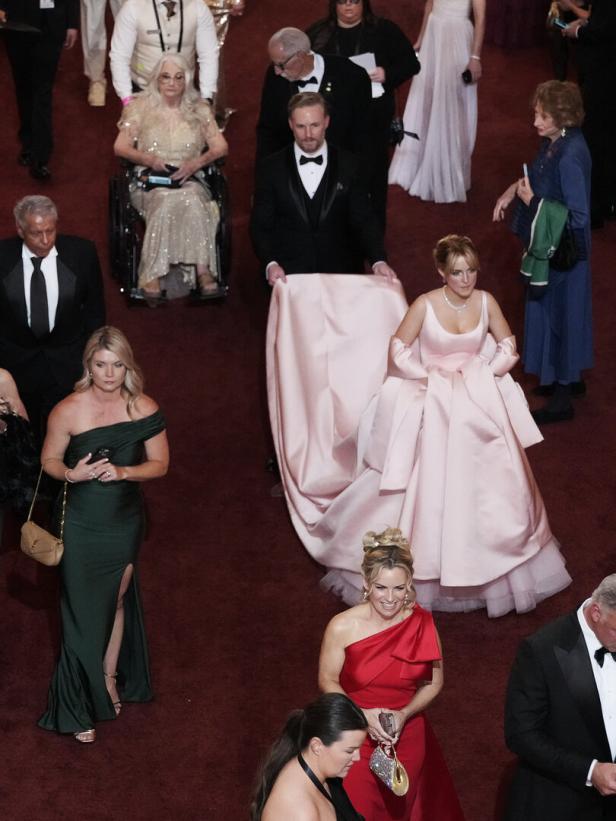Auf einem roten Teppich schreitet eine Frau in einem rosa Ballkleid, begleitet von einem Mann im Smoking.