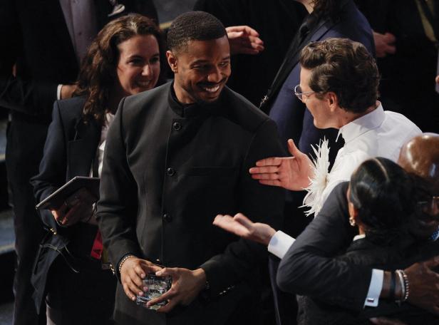 Michael B. Jordan lächelt mit einem Drink in der Hand, umgeben von anderen Personen.