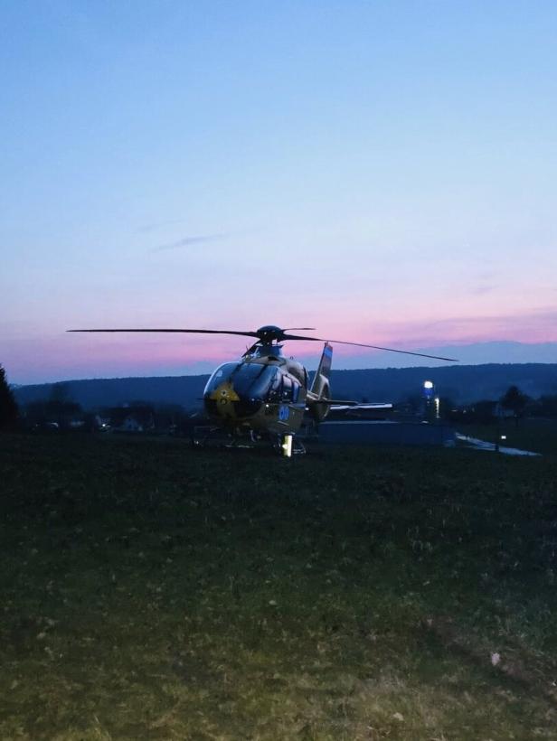 Ein gelber Rettungshubschrauber steht auf einer Wiese bei Sonnenuntergang, im Hintergrund sind Hügel und Häuser zu sehen.