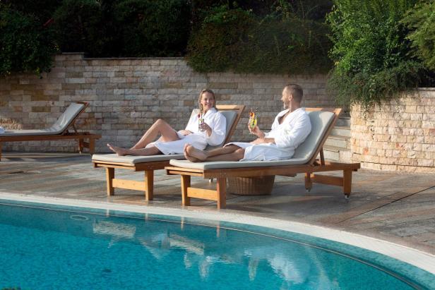Zwei Personen im Bademantel entspannen mit Getränken auf Liegen am Pool im Freien.