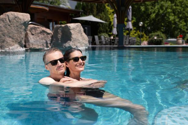 Paar entspannt gemeinsam im Außenpool einer Thermenanlage bei sonnigem Wetter.