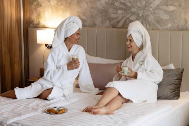 Zwei Frauen im Bademantel genießen Getränke und Gebäck entspannt auf einem Hotelbett.
