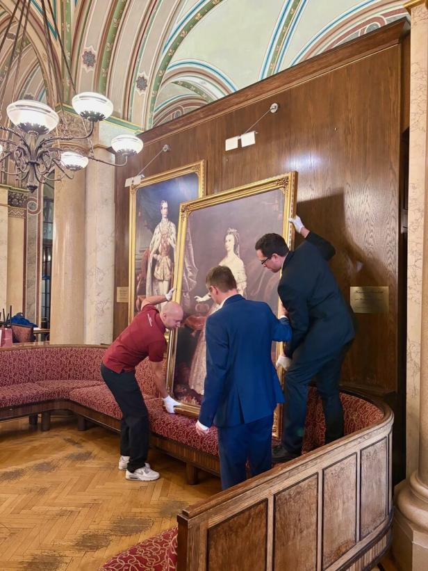 Drei Männer hängen ein großes, gerahmtes Porträt in einem prunkvollen, historischen Raum auf.