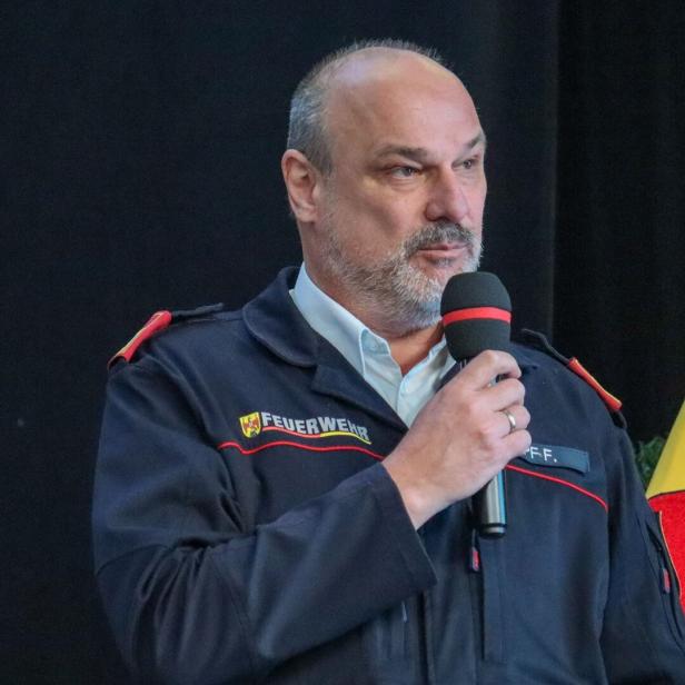 Ein Mann in Feuerwehruniform spricht mit einem Mikrofon in der Hand vor dunklem Hintergrund.