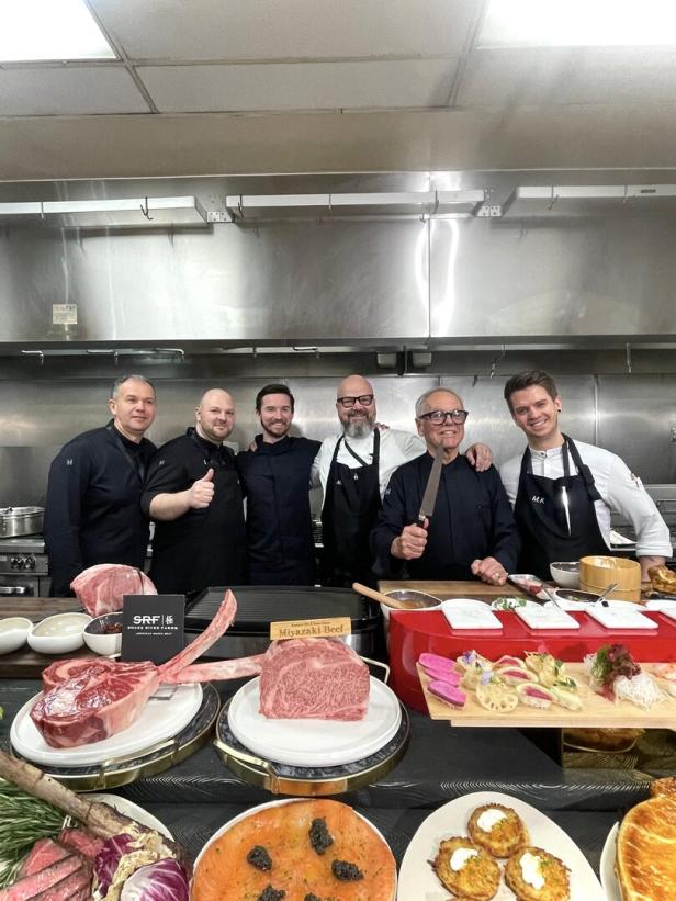 Sechs Köche stehen lächelnd hinter einer Theke mit rohem Fleisch, Sushi und weiteren Speisen in einer Restaurantküche.
