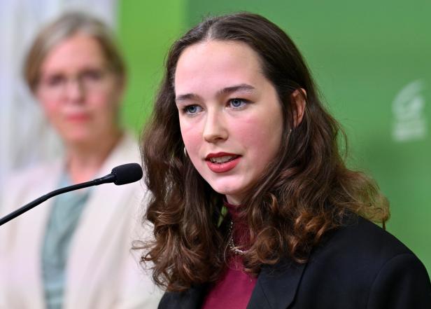 Junge Frau spricht am Mikrofon, im Hintergrund unscharf eine weitere Person vor grünem Hintergrund.