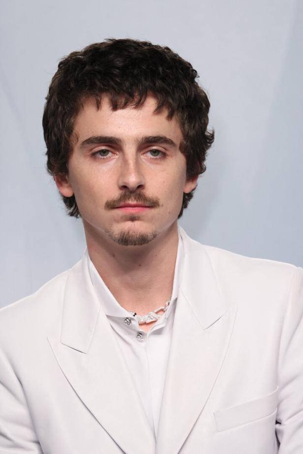 Timothée Chalamet mit dunklen, lockigen Haaren und Schnurrbart trägt einen weißen Anzug und eine auffällige Halskette.