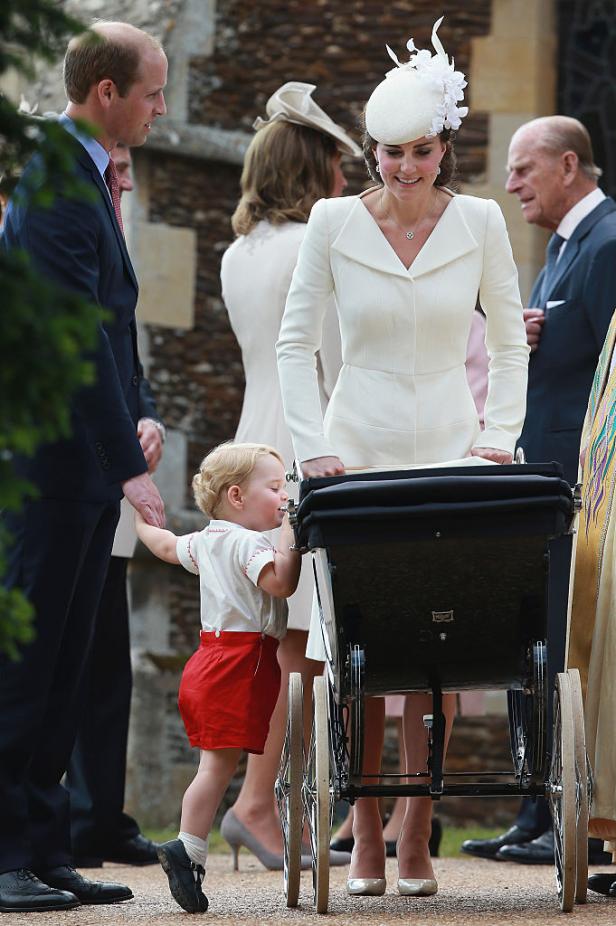 Prinz William, Prinzessin Kate und der kleine Prinz George am Tag von Prinzessin Charlottes Taufe (5. Juli 2015). Kate schiebt eleganz gekleidet einen Kinderwagen, George schaut auf Zehenspitzen stehend hinein.