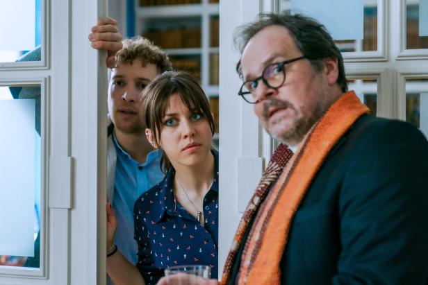 Eine Szene aus "So haben wir dich nicht erzogen": Thomas Mraz mit Brille und Schal blickt überrascht, während Alina Schaller und Julian Pichler durch eine Tür schauen.